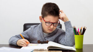 A young boy looked frustrated while doing homework at a desk, holding a pencil with study materials and colored pencils nearby.